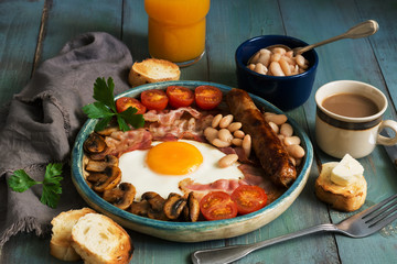 A traditional full English breakfast with fried egg, sausage, mushrooms, beans, bacon and tomatoes on a rustic wooden green table.