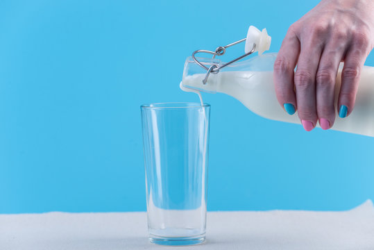 Woman's hand pours fresh milk from a glass bottle into a glass on a blue background. Healthy dairy products with calcium