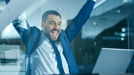 In the Office Businessman Sitting at the Desk Using Laptop Finishes Project and Wins Big. Makes Successful Gestures Raises Arms in Celebration.
