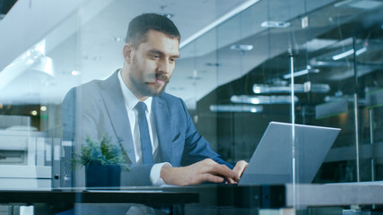 Confident Handsome Businessman Works on a Laptop at His Desk. Stylish Man in Modern Glass Office.