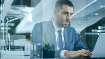Confident Handsome Businessman Works on a Laptop at His Desk. Stylish Man in Modern Glass Office.