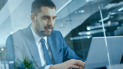 Confident Handsome Businessman Works on a Laptop at His Desk. Stylish Man in Modern Glass Office.