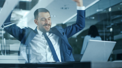 Handsome Businessman Works on a Laptop at His Desk, He Finished Job, Broke the Jackpot, Won Big and Is Happy and Celebrates His Success, Shows His Muscle. Stylish Man in Modern Glass Office.