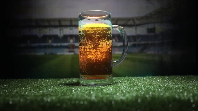 Creative concept. Pint of fresh beer on grass with blurred stadium on background with fog and light or Glass of lager beer ready for drink. Copy space. Selective focus