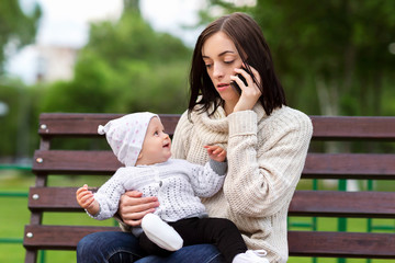 Busy upset mother is sitting with baby girl at a bench and talking by cellphone at park background.