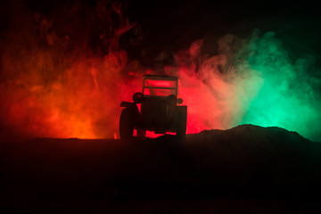 Silhouette of old vintage car in dark foggy toned background with glowing lights in low light.