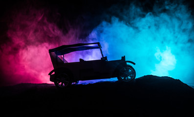 Obraz premium Silhouette of old vintage car in dark foggy toned background with glowing lights in low light.