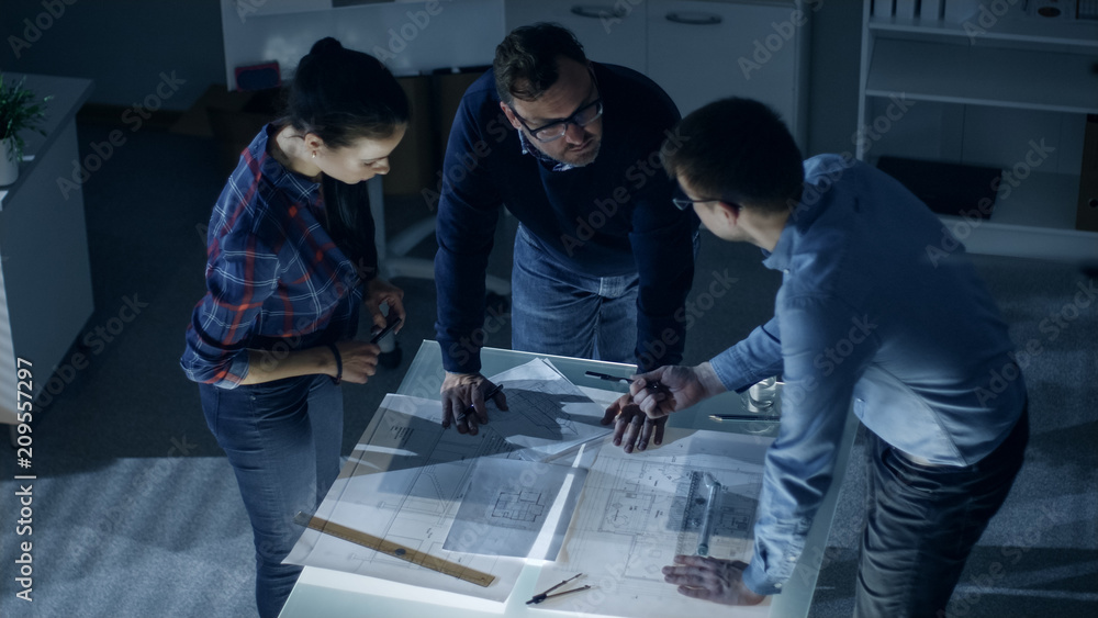 Poster late at night team of design engineers work with drafts and documents on illuminated table.