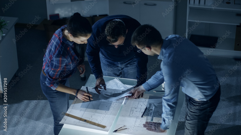 Poster late at night team of design engineers work with drafts and documents on illuminated table.