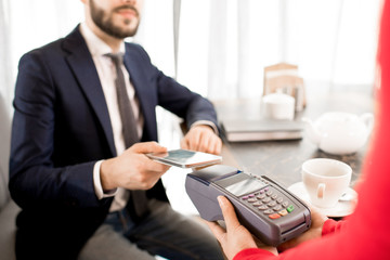 Close-up of modern businessman using contactless payment while performing transaction with smartphone in restaurant