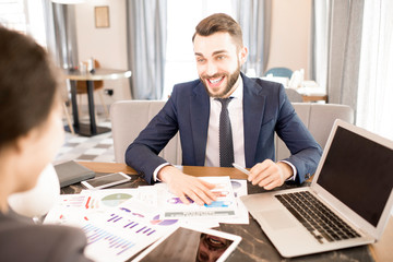 Cheerful excited bearded young businessman in formal suit laughing while discussing statistics with colleague in cafe, business analysts working together