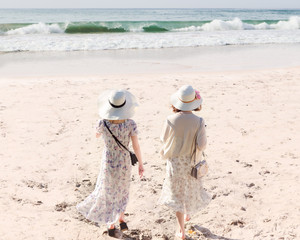 Rear view of two young women in long dresses and hats walking along sandy beach meet sea water....