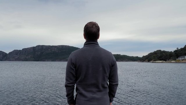 Medium Shot Back view. Male tourist meditatively peering at the beautiful landscape. Amazing pond, lake or sea with calm smooth water surface. High mountains covered dense green forest, clear blue sky