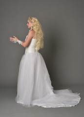 full length portrait of blonde  girl wearing white fantasy gown. standing pose in side profile, grey studio background.