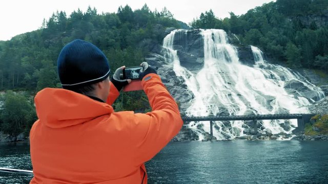 Medium Shot. Back side view. Natural cascade in Norway. Amazing island, high mountains, covered dense green forest. Beautiful nature, journey or cruise. Quiet clean pond. Traveler in warm jacket.