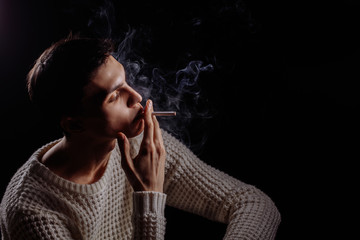 Thougtful young man, wearing warm knitted white sweater smoking sigarette, thinking on dark...