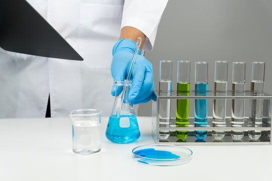 Laboratory Scene, The Scientist Holding The Clipboard And Blue Liquid Flask With Glass Stirring Rod, Test Tubes In The Rack, Watch Glass And Beaker On The Table