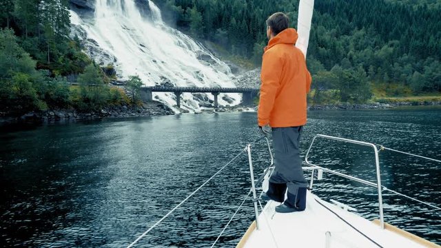 Natural cascade in Norway. Amazing island, high mountains, covered dense green forest. Beautiful nature, journey or cruise. Quiet clean pond. Traveler in warm jacket. Low Angle. Full Shot, side view