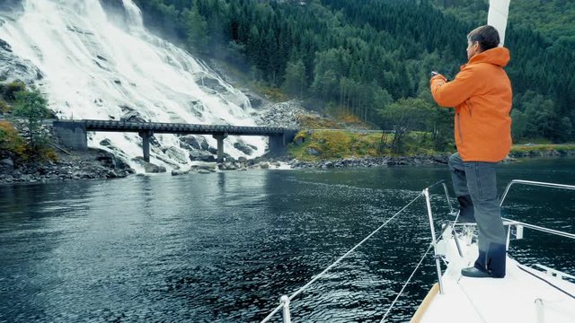 Tourist making a photo of natural cascade in Norway. Amazing island, high mountains, covered dense green forest. Beautiful nature, journey or cruise. Traveler in warm jacket. Low Angle. Full Shot