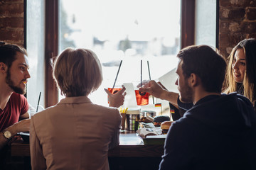 Back view of cheerful company of young happy gyus and girls talking while sitting at bar counter in a modern urban cafe near the window at daytime.