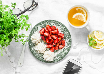 Breakfast or snack table - fresh strawberries, meringue, green tea with lemon, smartphone and headphones. Cozy home rest break still life on a light background, top view