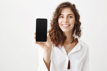Indoor portrait of charming young woman with curly hair smiling broadly while holding smartphone, advertising gadget while standing over gray background in trendy blouse. great present for valentines