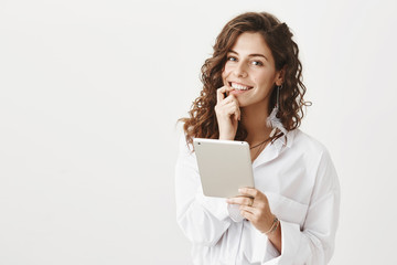 Portrait of charming woman with curly hair biting finger and smiling sensually at camera while holding tablet and standing over gray background. Boyfriend making breakfast while girl plays in gadget