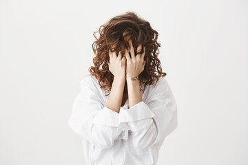 Studio portrait of tired or bothered woman hiding face in her curly hair and holding hands on it, expressing exhaustion or just being childish, standing over gray background. I am not seeing you