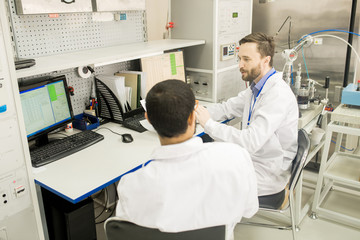 Two bearded lab technicians analyzing statistic data on computer connected to control cabinet while gathered together at modern measuring equipment factory