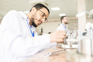 Fototapeta premium Profile view of concentrated Arabian engineer wearing lab coat sitting at desk and assembling pressure sensor, interior of measuring device factory on background
