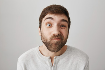Indoor shot of attractive funny bearded guy squinting and making faces at camera, being in good mood while standing over gray background. Man mimics as if he looking through peephole