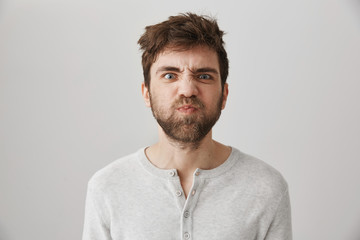 Obraz premium Portrait of funny weird guy with messy hair and beard making faces, puckering eyebrows and sulking, standing over gray background with offended or irritated expression, losing his temper