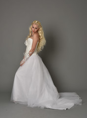 full length portrait of blonde  girl wearing white fantasy gown. standing pose in side profile, grey studio background.