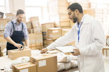Profile view of bearded Arabian inspector wearing lab coat holding clipboard in hand while carrying...