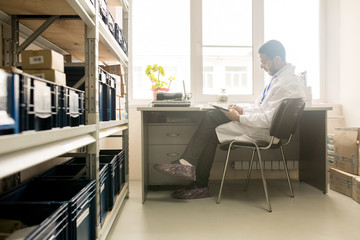 Profile view of concentrated bearded technician wearing white coat sitting at desk and taking...