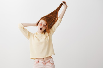 Everyone is jealous of her beautiful hair. Portrait of playful expressive young redhead in fashionable clothes and pink glasses pulling hair up with hand and gasping from excitement over gray wall