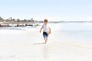 Fototapeta na wymiar Happy three years old child boy in yellow hat running at white sand and jumping in the waves during summer vacation on tropical beach near blue sea. Smiling and cute toddler kid at the sea