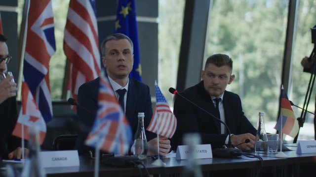 Adult male representative of America telling speech into microphone while sitting at table on international summit with other members