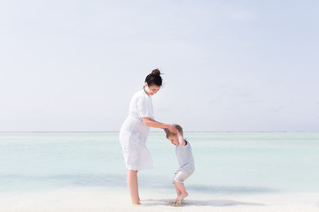 Fototapeta na wymiar Little child with mom on the beach. Kid boy playing with his mother, hugs and holds his mother's hand and smiles. Young woman in white dress resting with her toddler son at the seaside.