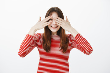 Studio portrait of happy excited girl seeing awesome surprise. Impatient charming woman in casual outfit, covering eyes with palms and peeking through fingers, smiling broadly over gray wall