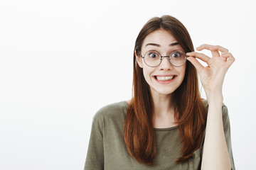 Waist-up portrait of good-looking funny woman in stylish glasses, holding rim with fingers and smiling with strange expression, fooling around, making faces while standing over gray background