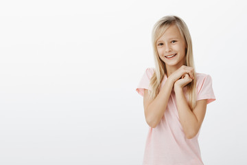 Indoor shot of adorable joyful young female child with blond hair, holding hands together near face...