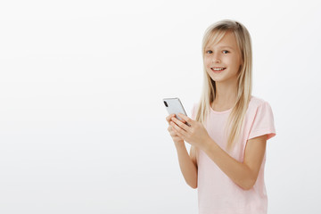 Fototapeta premium Carefree adorable joyful girl with blond hair wearing casual pink t-shirt, holding smartphone and gazing at camera with broad smile, feeling boost of happiness receiving like under new photo
