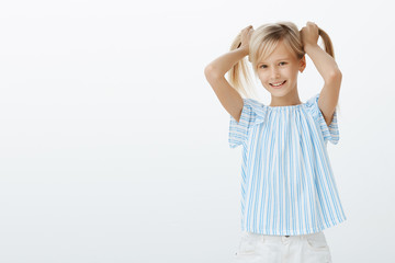 Girl showing friends her new earrings. Joyful pleased little girl with blond hair, lifting hair up and smiling joyfully while advertising cute new accessorize, standing against gray background