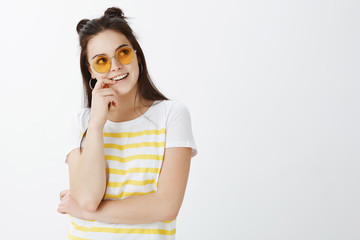 Waist-up shot of stylish creative young european female in trendy sunglasses, looking right with intrigued and joyful expression, smiling and biting fingernail while thinking and having idea in mind