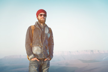 Naklejka premium Hipster young man with beard and mustache wearing sunglasses posing against the background of mountains