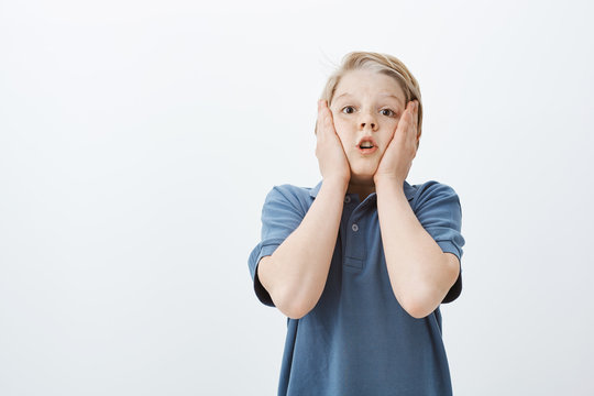 Studio shot of impressed amazed cute male child in blue t-shirt, holding palms on cheeks, holding breath and gasping from shock and surprise, staring at camera and tilting head backwards