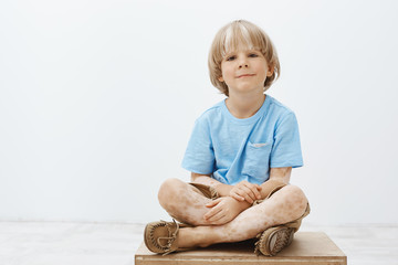 Indoor shot of cute happy blond child with positive smile sitting with crossed hands, having...