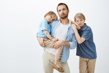 Bored skeptic european dad in casual outfit holding little kid with vitiligo, rolling eyelids from annoyance or tiredness, older son leaning on father shoulder and gazing at camera with indifference