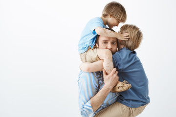 Close-up shot of tired worried father holding little son on shoulders while older son hanging on chest and hugging dad, staring at camera with concerned expression, playing with energetic kids